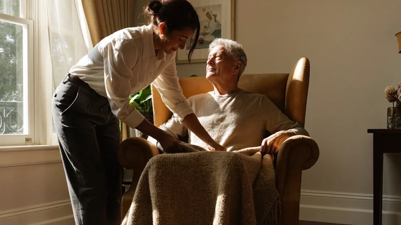 Senior im Sessel, Pflegekraft ordnet eine Decke in sonnigem Wohnzimmer, Illustration
